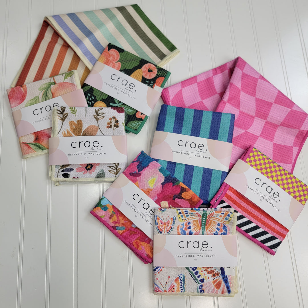 Crae Home Washcloths and Towels by Cosmos + Clover Boutique are arranged on a white surface, featuring colorful stripes, florals, and geometric patterns in vibrant hues.