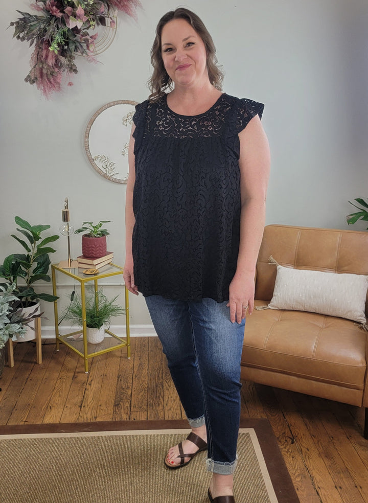 A woman with wavy brown hair smiles in a cozy living room, wearing the Heimish Eden Lace Top with blue jeans and sandals. The background features plants, a tan couch, and wall decor.