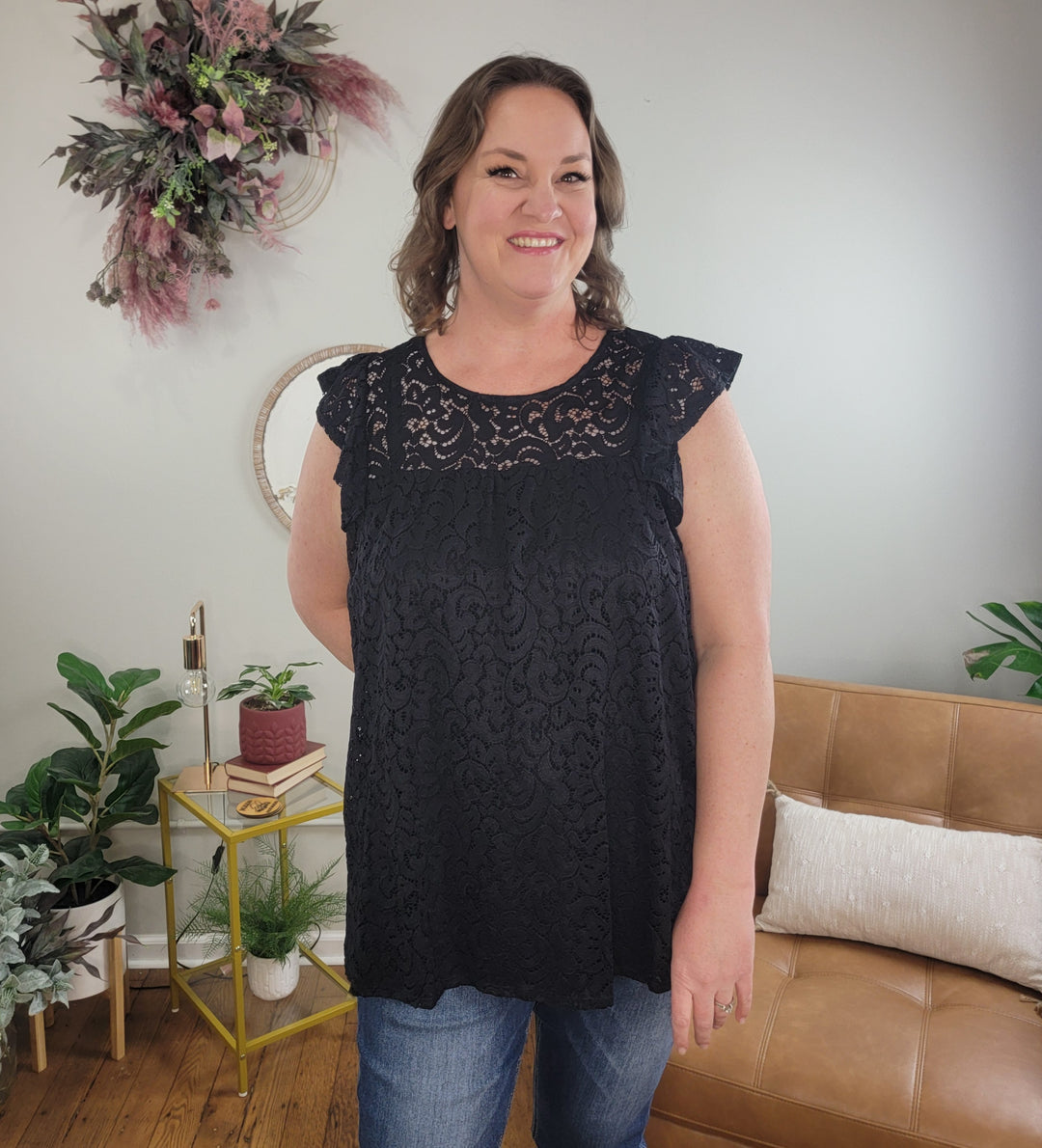 A woman wearing the Heimish Eden Lace Top and blue jeans stands smiling in a cozy, modern living room with plants, a tan sofa, and decorative wall art in the background.
