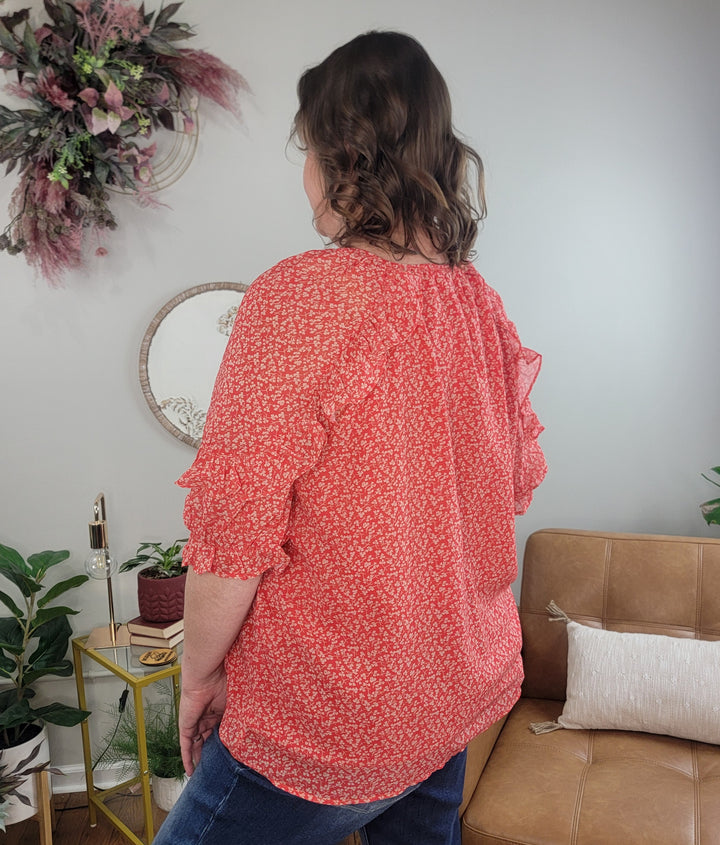 A person with wavy brown hair, wearing the Hayden Chase Ruffle Top and jeans, stands with their back to the camera in a cozy room featuring plants, a tan sofa, a gold side table, and stylish wall decor.