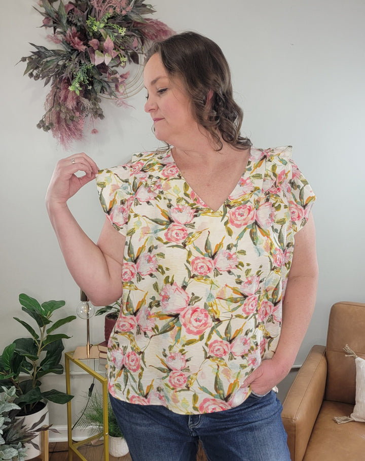 A woman in the Jupiter Floral Top by Umgee and jeans stands indoors, gently touching her shoulder. Potted plants, a tan armchair, and a floral wall decoration are behind her.