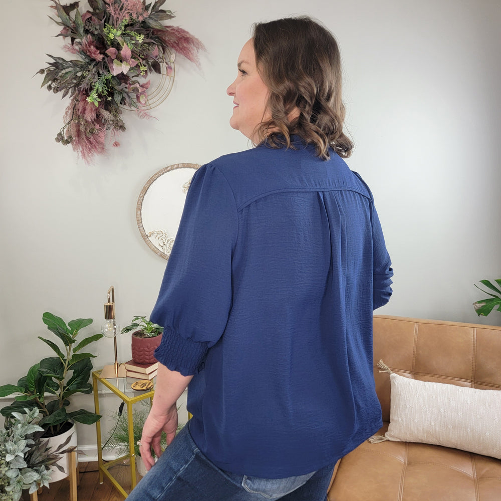 A woman with wavy brown hair, wearing the Umgee Remington Navy Top and jeans, stands with her back to the camera in a cozy room featuring plants, a beige couch, and a wall-mounted floral arrangement.