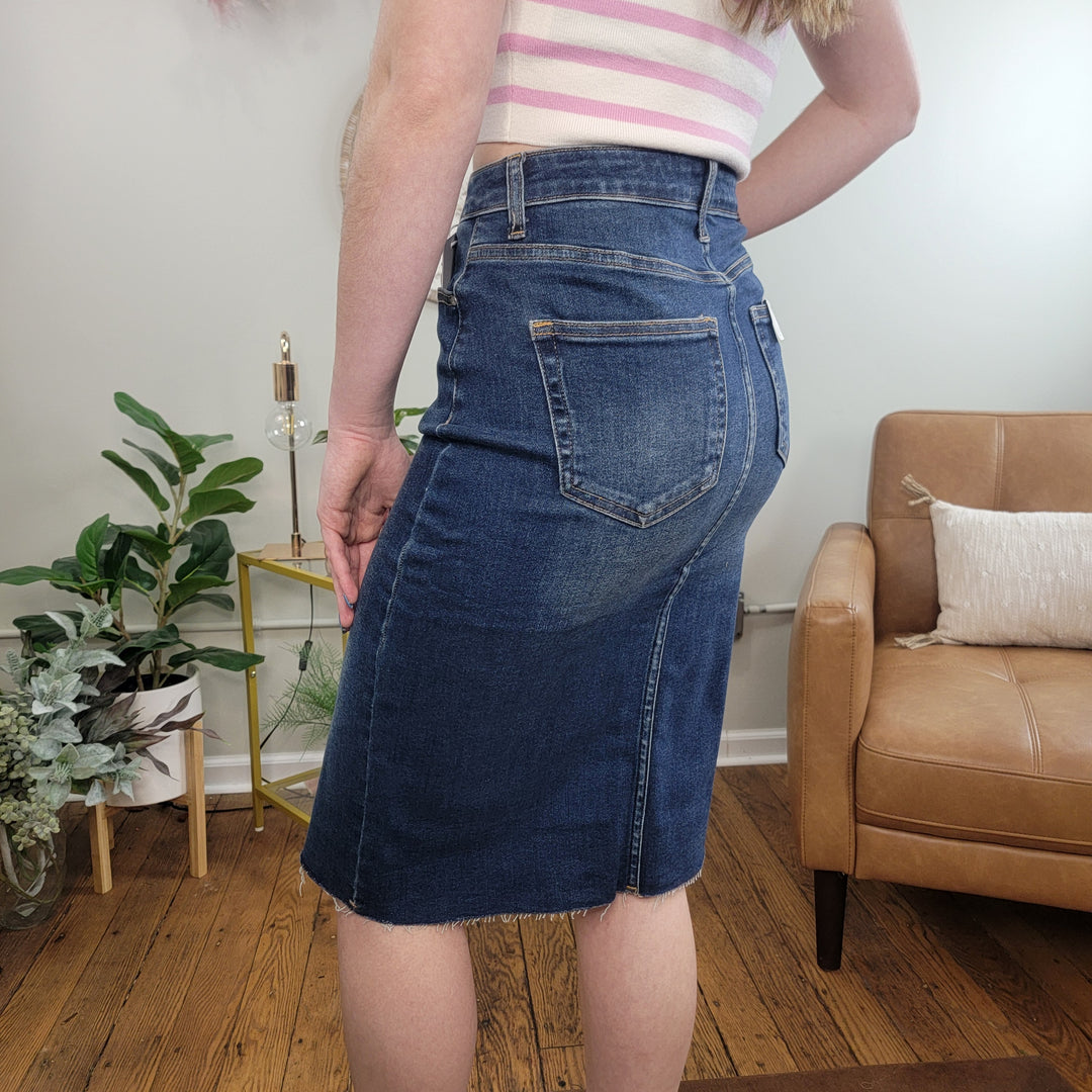 A person is seen indoors near a tan armchair and green plants, wearing the Risen Mickey Denim Midi Skirt with a sleeveless striped top. The image highlights the back and side view of the dark wash skirt on a wooden floor.