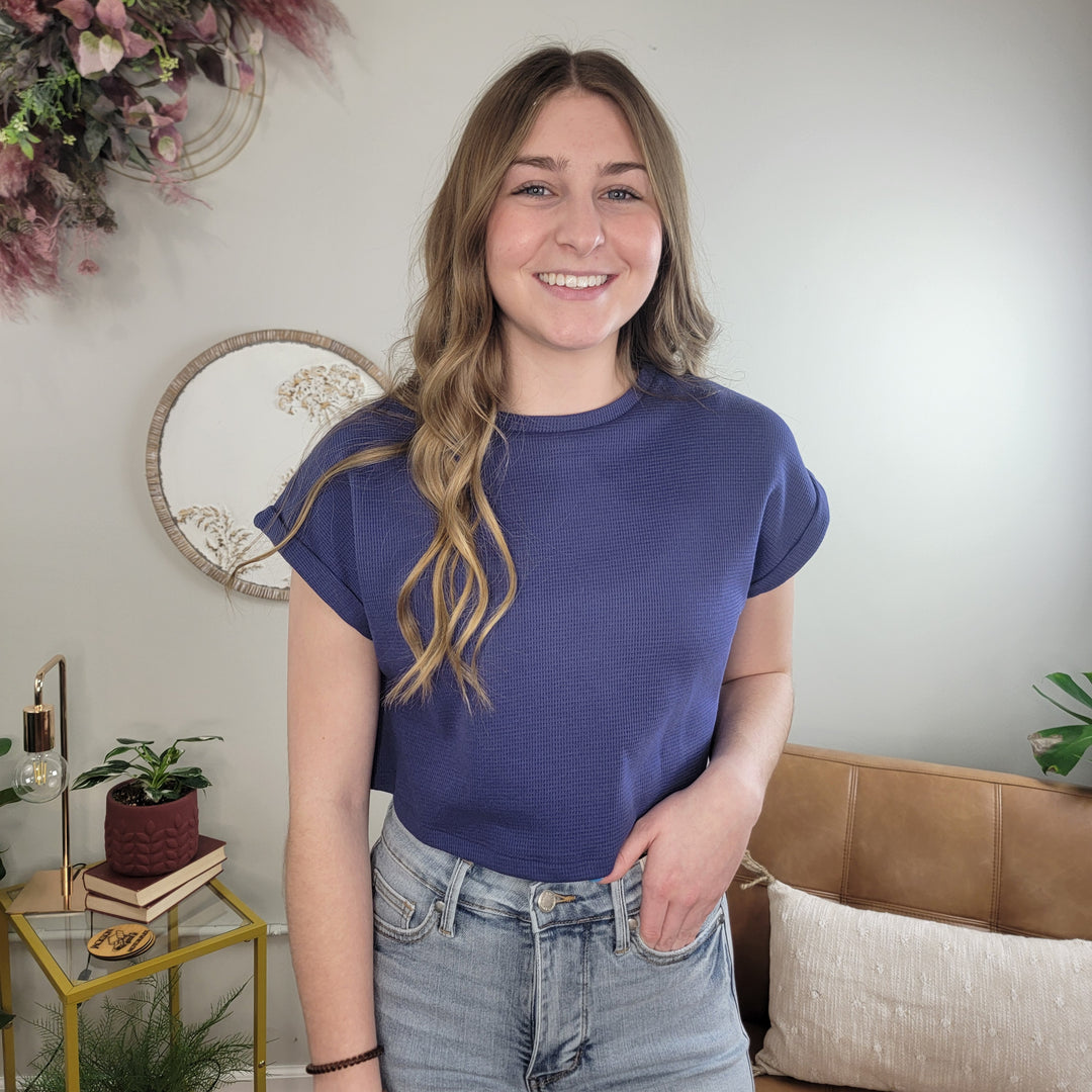 A young woman with wavy light brown hair, wearing the HYFVE, Inc Tinsley Crop Tee in dusty blue and light jeans, stands smiling in a cozy living room decorated with plants, books, and wall art.