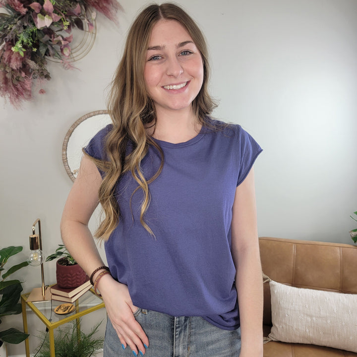A young woman with long, wavy hair smiles indoors wearing the HYFVE, Inc. Meriah Tee in purple and jeans. Behind her are a brown couch, small table, books, plants, and wall decor.