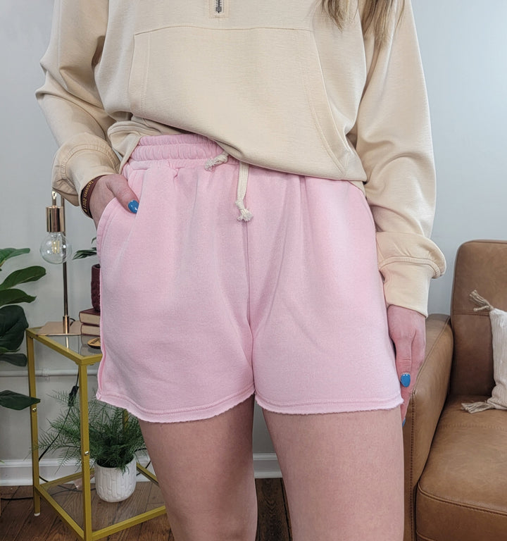 Someone stands indoors with one hand in their pocket, wearing cream and light pink Mabel Fleece Shorts by HYFVE, Inc. A small yellow side table with a plant and lamp is visible in the background.