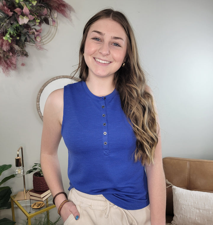 A young woman with long wavy hair wears the Adina Top by HYFVE, Inc—a royal blue ribbed knit tank—paired with beige pants. She smiles indoors, with decor, a sofa, and green plants in the background.