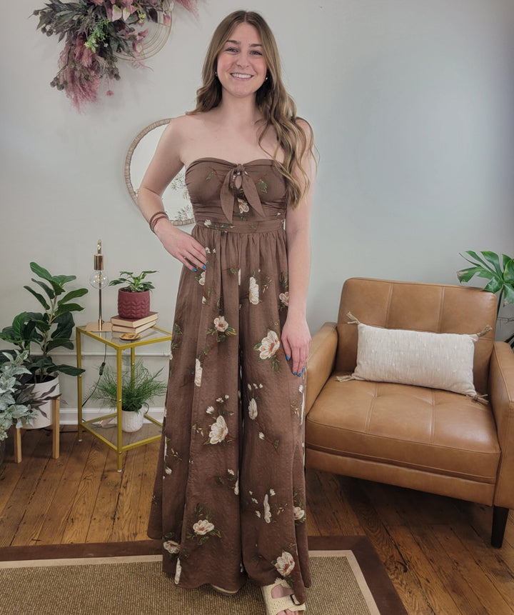 A woman smiles in a cozy living room, wearing the HYFVE, Inc Poppy Floral Jumpsuit. Behind her are a tan armchair, green plants, a small table, and wall decor.