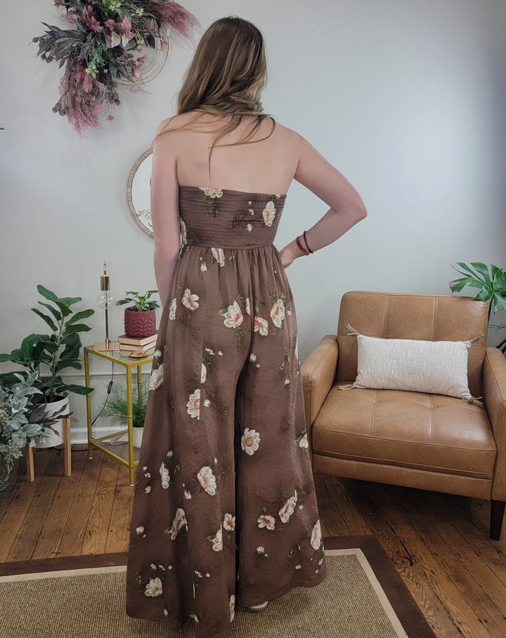 A woman with long hair stands indoors on a rug near a tan armchair, wearing the cute and stylish Poppy Floral Jumpsuit by HYFVE, Inc—perfect for spring and summer events amidst plants and decorative accents.
