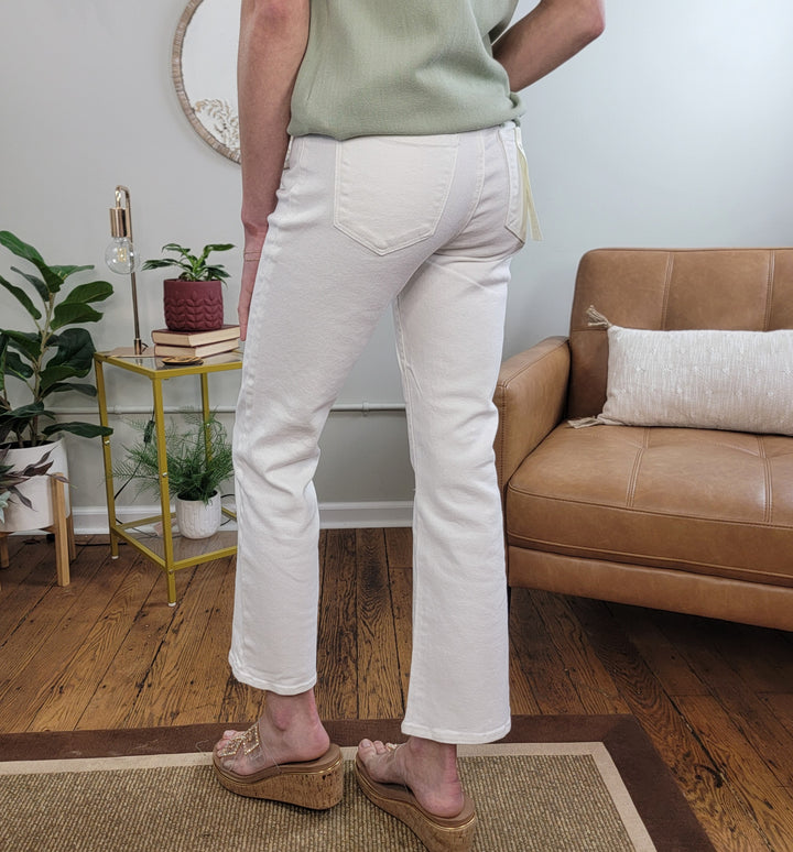 A person stands indoors on a rug, wearing Risen Acacia Tummy Control Crop Bootcut Jeans in white, a light green top, and beige wedge sandals. Behind them are a brown leather chair, gold side table with plants and books, and a white wall with round mirror.