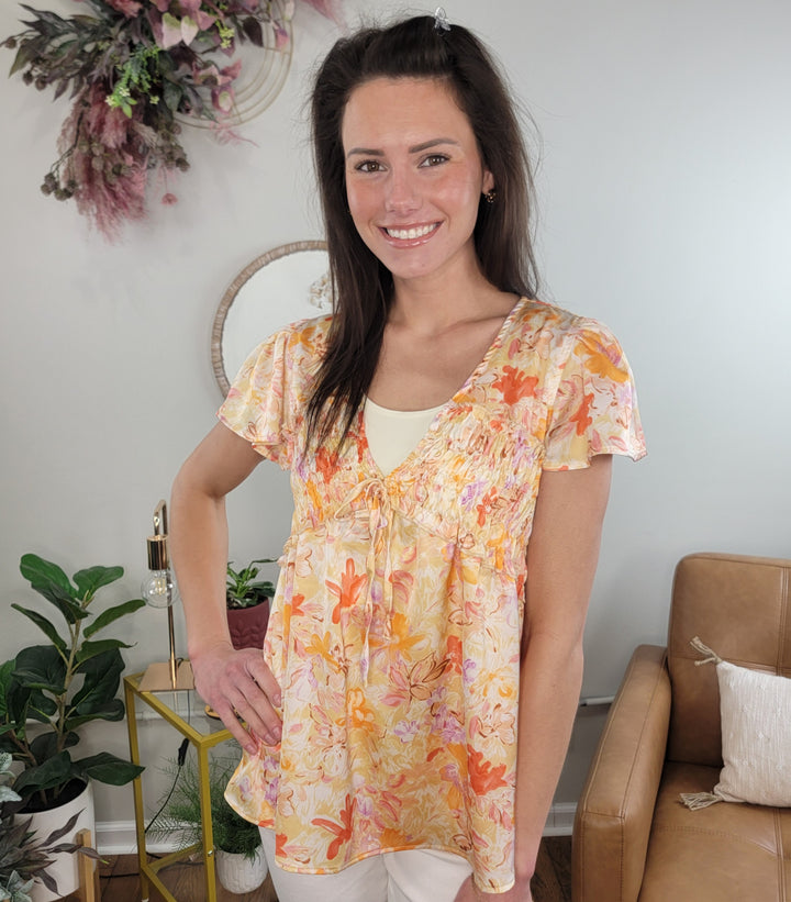 A woman with long brown hair smiles in a cozy room, wearing the Doe &amp; Rae Amara Floral Print Top and light pants, surrounded by plants, a leather chair, and stylish wall decor.