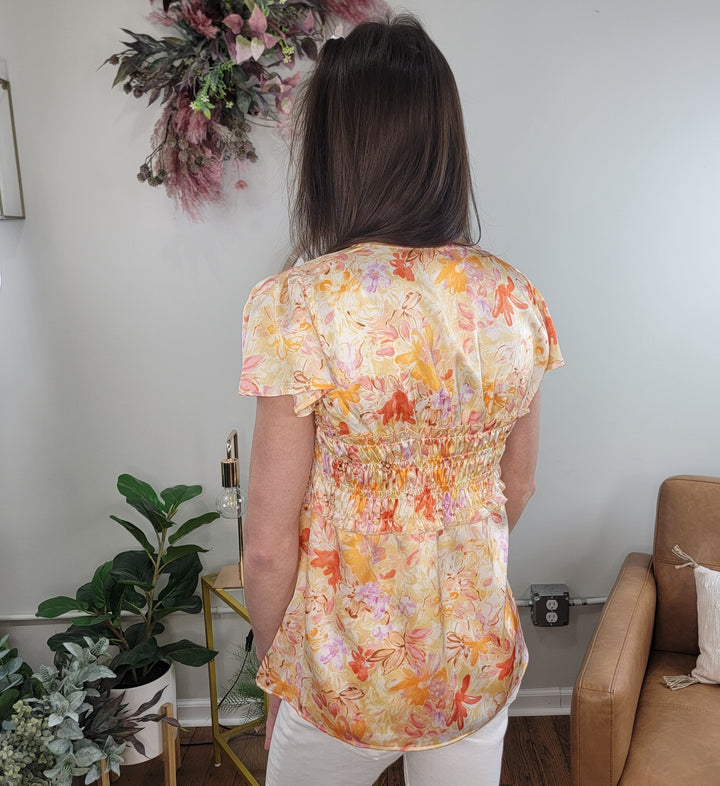 A woman with long brown hair stands facing away in a cozy living room filled with green plants and a brown chair, wearing the Amara Floral Print Top by Doe &amp; Rae in orange and pink, paired with white pants.