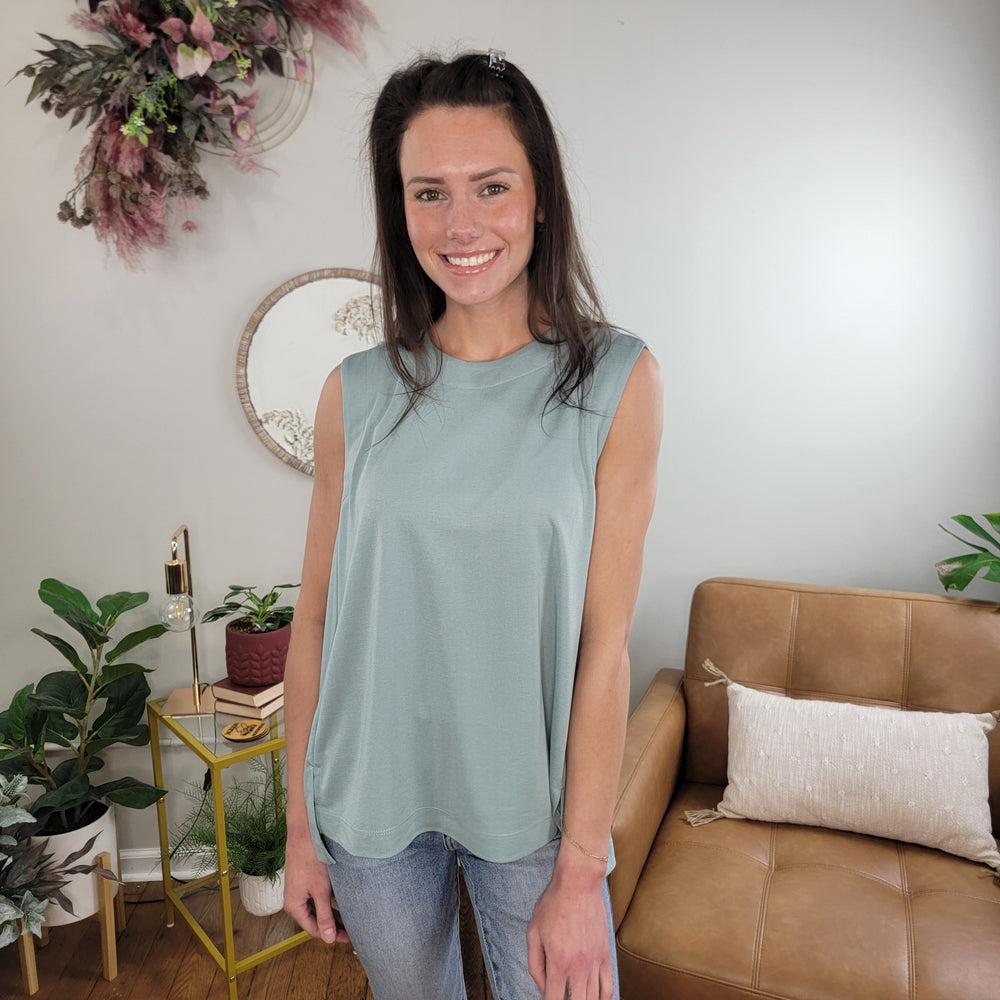A woman with long brown hair, wearing the Doe & Rae Beatrix Sleeveless Top in dusty blue and jeans, smiles in a cozy living room with plants, a tan sofa, and decorative wall art.