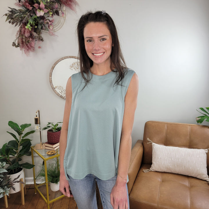 A woman with long brown hair, wearing the Doe & Rae Beatrix Sleeveless Top in dusty blue and jeans, smiles in a cozy living room with plants, a tan sofa, and decorative wall art.