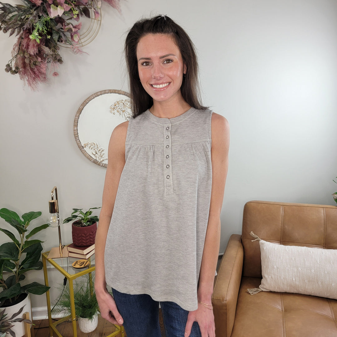 A woman with long brown hair smiles in a cozy living room, wearing the Doe & Rae Lillee Babydoll Tank and jeans, with plants, wall decor, and a tan couch in the background.