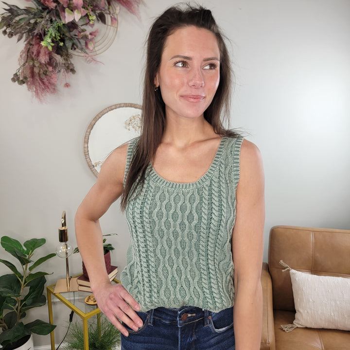 A woman indoors smiles slightly while wearing the Staccato Vicky Cable Knit Tank Top in jade and blue jeans. Behind her are plants, a circular wall décor, and a tan sofa with white pillows.