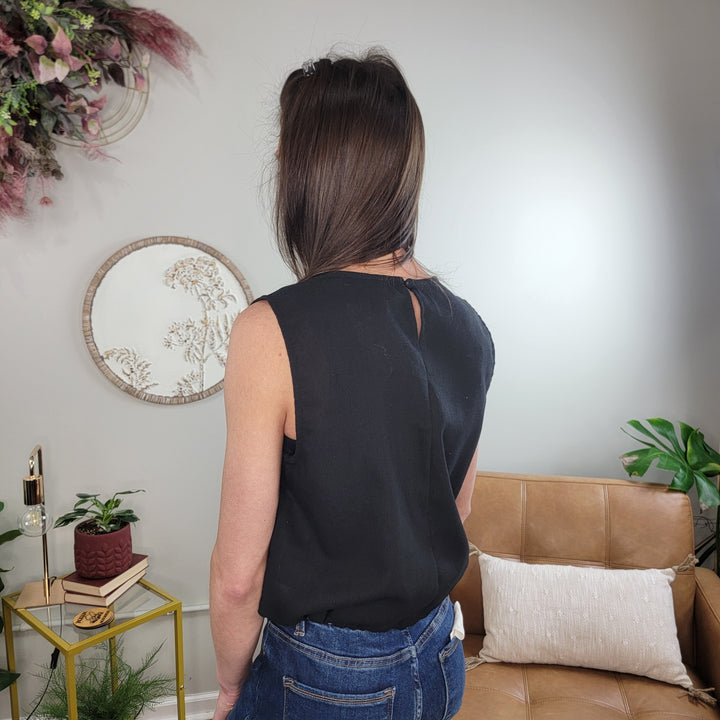 A woman with straight brown hair stands indoors with her back to the camera, wearing the HYFVE, Inc Reign Top—a sleeveless black crew neck—paired with blue jeans. The room features plants, a tan couch, and neutral decor.