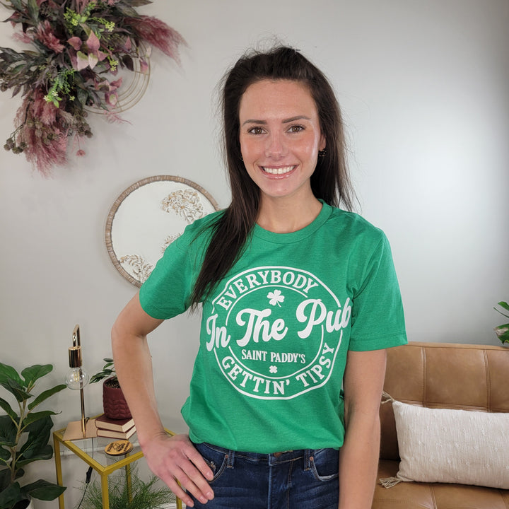 A woman with long brown hair smiles in a plant-filled living room, wearing the Tipsy in the Pub Graphic Tee by Cosmos + Clover Boutique—a comfy green shirt that adds festive flair to her relaxed look.