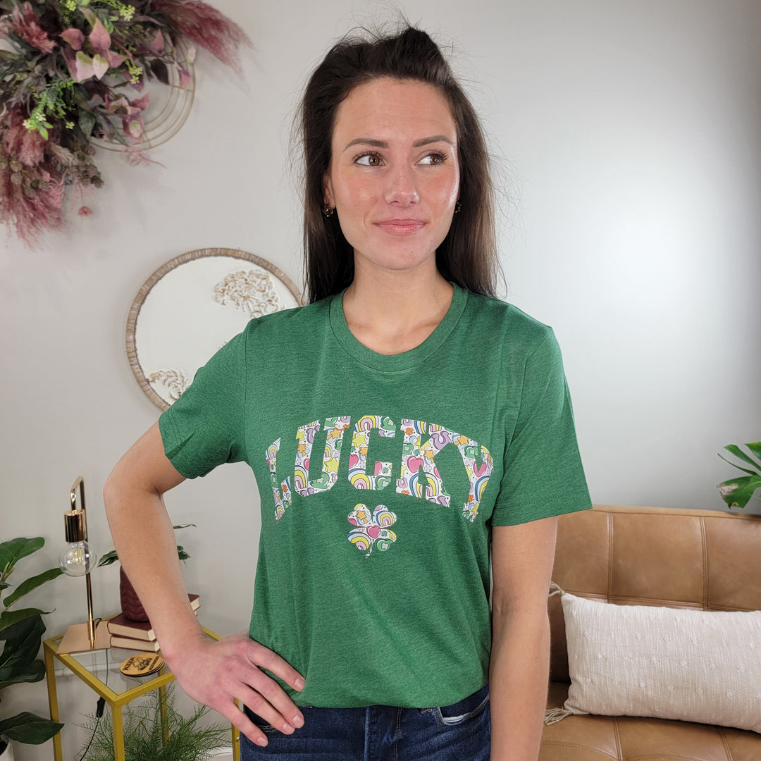 A woman with long brown hair stands indoors wearing the Lucky Charm Graphic Tee by Cosmos + Clover Boutique, featuring a colorful graphic. She smiles with one hand on her hip, surrounded by plants and stylish decor.