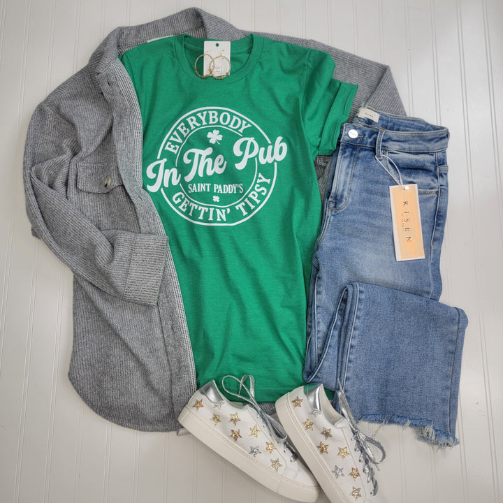 Flat lay: Tipsy in the Pub Graphic Tee by Cosmos + Clover Boutique, gray cardigan, light blue jeans with tag, and white sneakers with gold stars—all arranged on a light background for a festive Saint Patrick’s Day outfit.