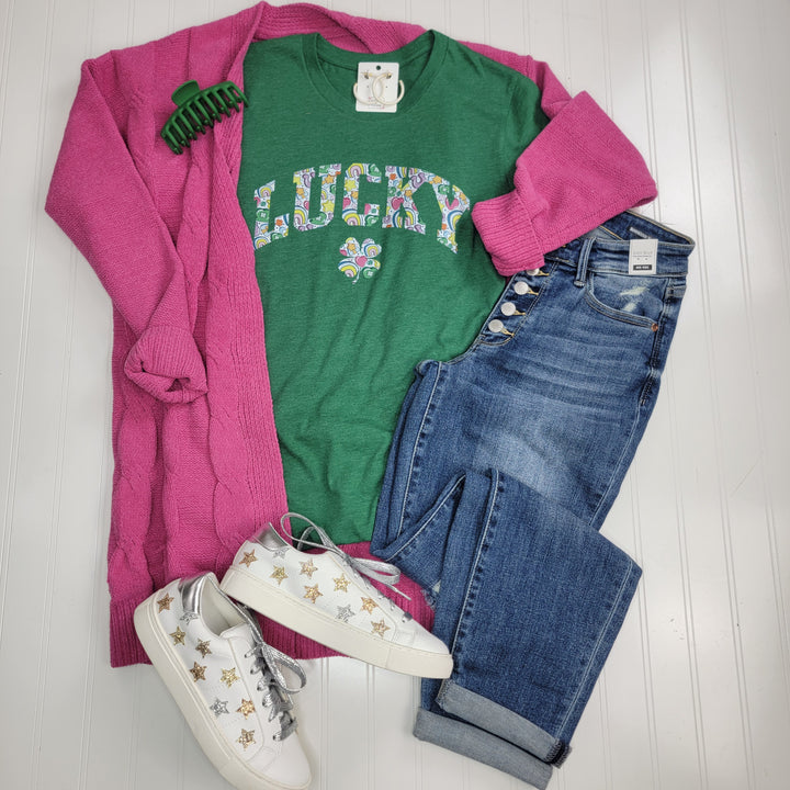 A casual outfit featuring the Lucky Charm Graphic Tee by Cosmos + Clover Boutique in heather green, paired with a pink cardigan, blue jeans, gold-starred white sneakers, a green hair clip, and white earrings on a white background.
