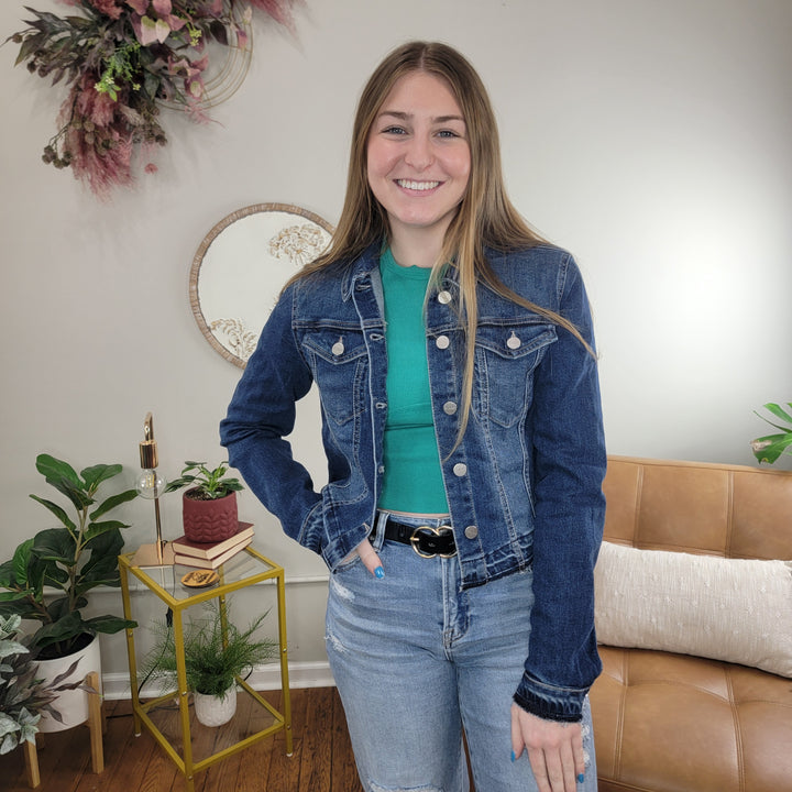 A smiling woman with long hair stands indoors next to a tan couch, wearing the Risen Kylie Jean Jacket over a green shirt and light blue jeans. Plants, a small table, and a round mirror decorate the background.