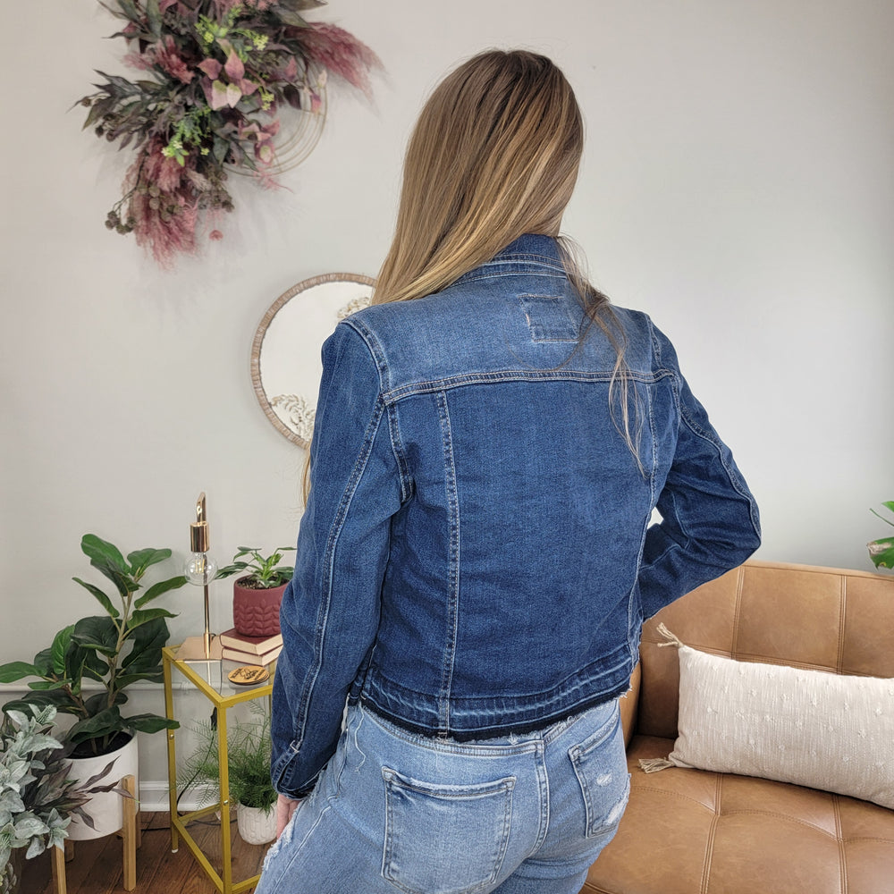 A woman with long, light brown hair stands with her back to the camera, wearing the Risen Kylie Jean Jacket and jeans in a cozy living room decorated with plants, a tan sofa, and wall art.