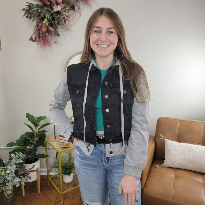 A young woman smiles indoors, wearing Risen’s Meadow Hooded Jean Jacket over a green top and ripped jeans. Behind her are plants, a tan sofa, and decorative wall art.