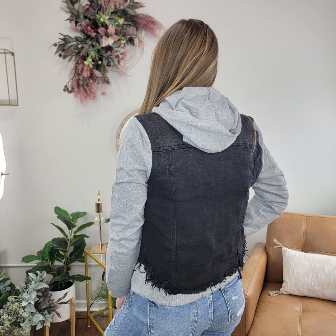 A woman with long hair faces away indoors, wearing the Risen Meadow Hooded Jean Jacket over blue jeans in a cozy room with plants, a tan couch, and floral wall decor.