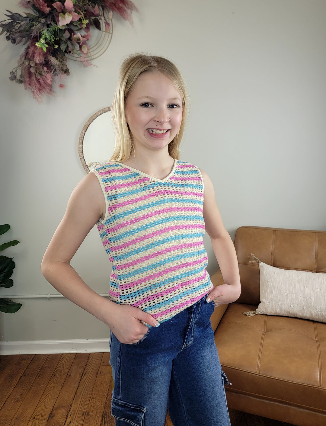 A smiling girl with blonde hair stands indoors with hands in her pockets, wearing the Hayden Girls Marty Open Knit Tank and blue jeans. A brown sofa and wall decor can be seen in the background.