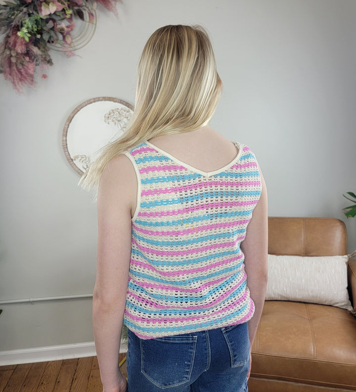 A girl with long blonde hair stands indoors facing away, wearing the Hayden Girls Marty Open Knit Tank and blue jeans. The room has a brown sofa, a plant, floral wall decor, and a round mirror.
