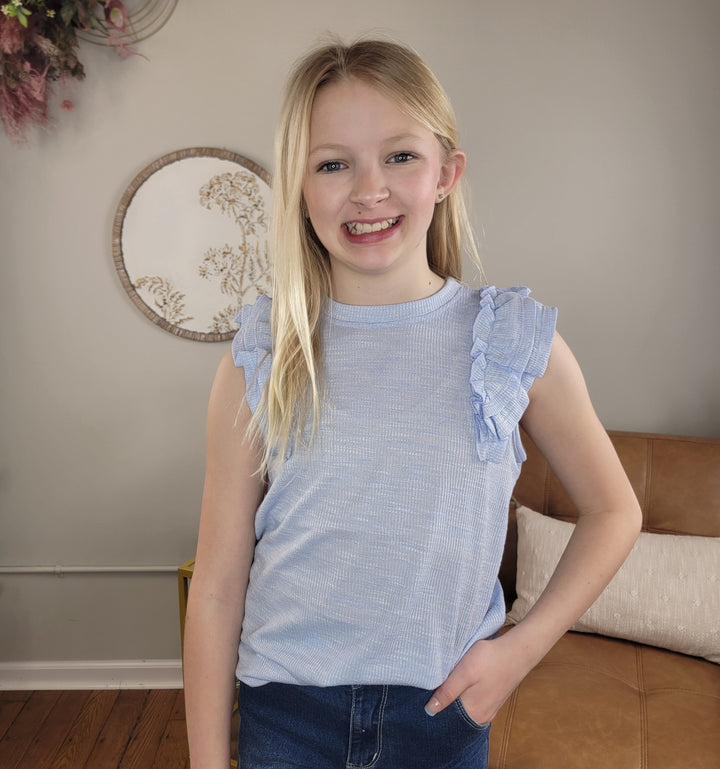 A young girl with long blonde hair smiles at the camera, wearing the ice blue Girls Jemma Top by Hayden and blue jeans. She stands indoors in a cozy room with a comfy couch and stylish wall decor behind her.