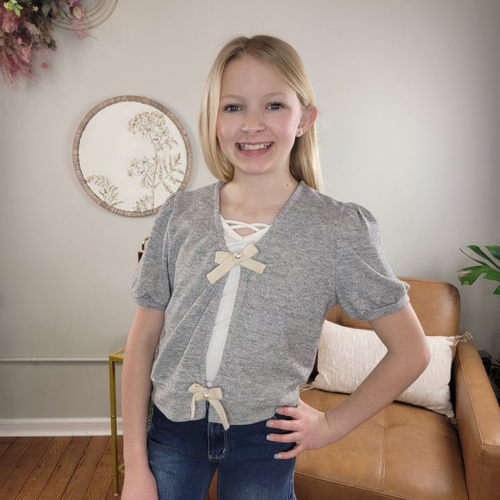 A smiling young girl with long blonde hair wears the Hayden Kids Zoe Sweater in heather grey over a white top and blue jeans. She stands in a cozy, well-lit room with neutral decor and a tan chair.