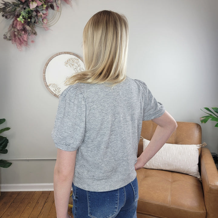 A woman with blonde hair models the Hayden Kids Zoe Sweater in heather grey and blue jeans, standing with her back to the camera in a neutrally decorated living room featuring a tan chair and greenery.