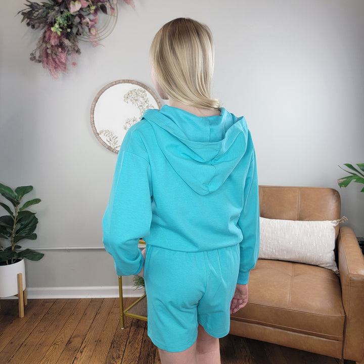 A person with blonde hair, seen from behind, wears the Cutie Patootie Girls Chloe Zip Up Hoodie in turquoise with matching shorts. They're standing in a cozy room with wooden floors, a tan couch, plants, and a floral round wall mirror.