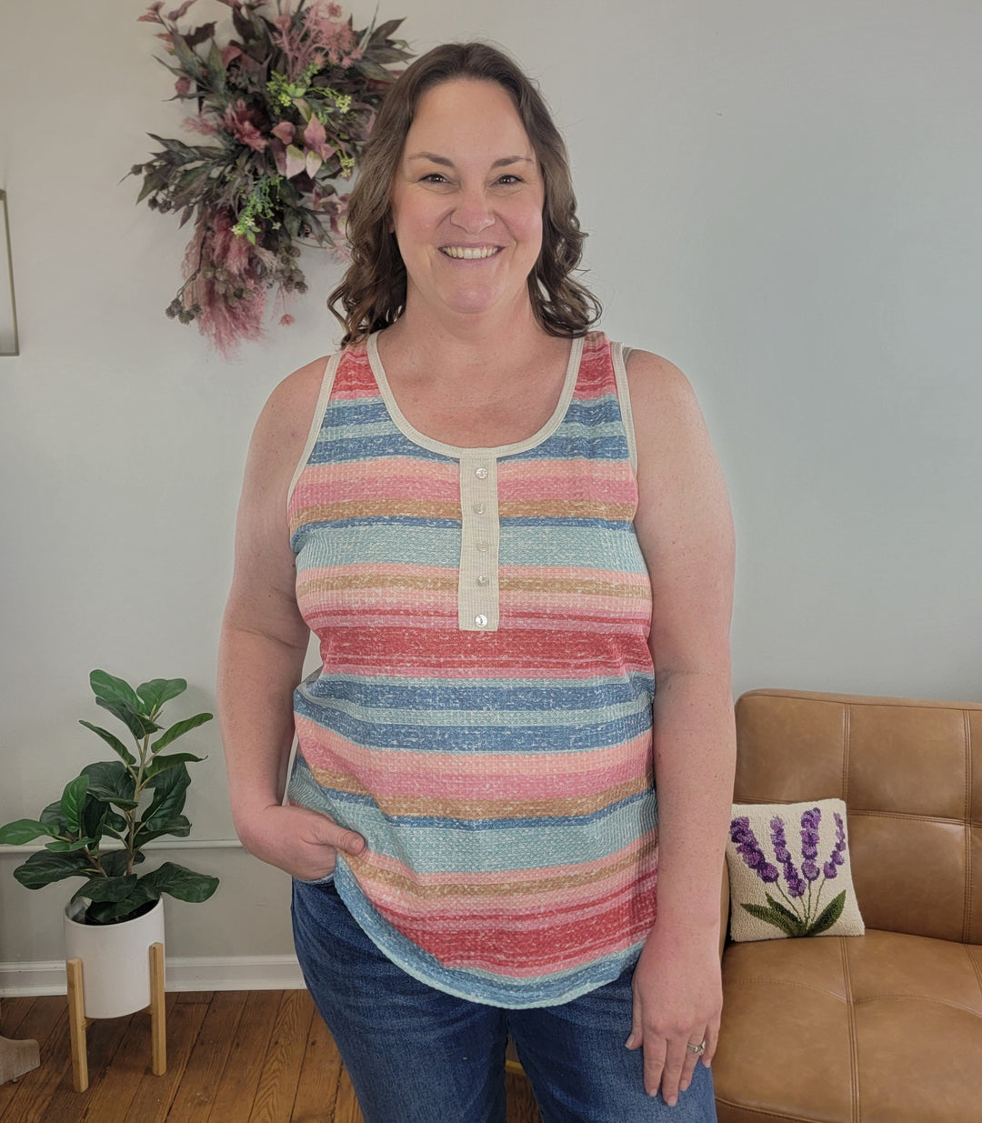 A woman with wavy brown hair smiles indoors, wearing the Heimish Camila Tank Top and jeans with a hand in her pocket. The background features plants, a floral wall wreath, and a tan sofa.