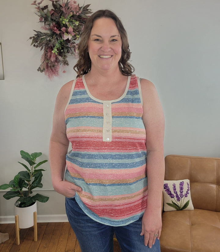 A woman with wavy brown hair smiles indoors, wearing the Heimish Camila Tank Top and jeans with a hand in her pocket. The background features plants, a floral wall wreath, and a tan sofa.