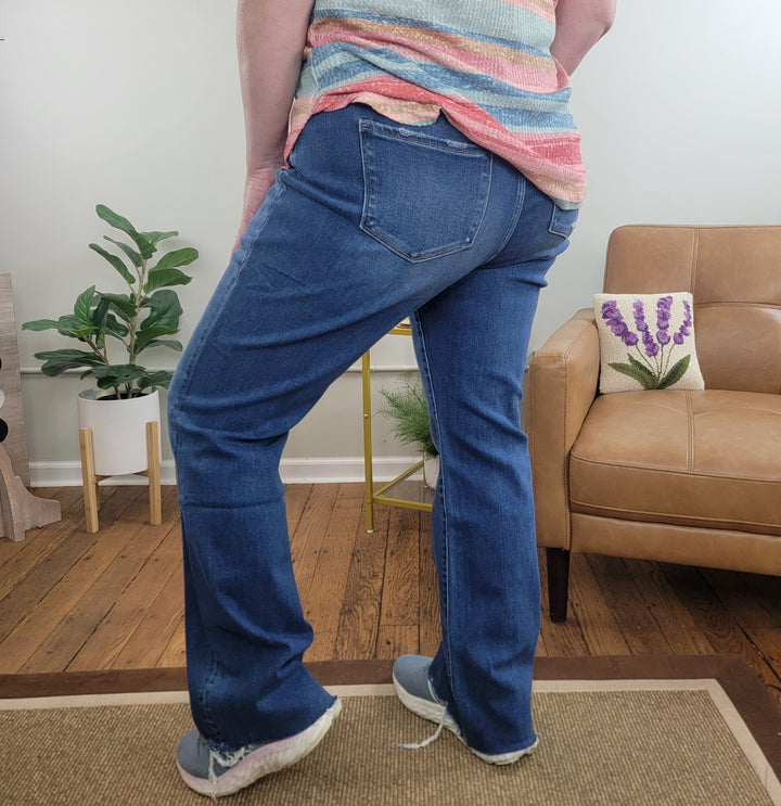 A person stands indoors facing away, wearing Lovervet Romi Tummy Control Bootcut Jeans with a striped top and gray shoes. The scene features a tan armchair, plant, wooden floors, and a small rug.