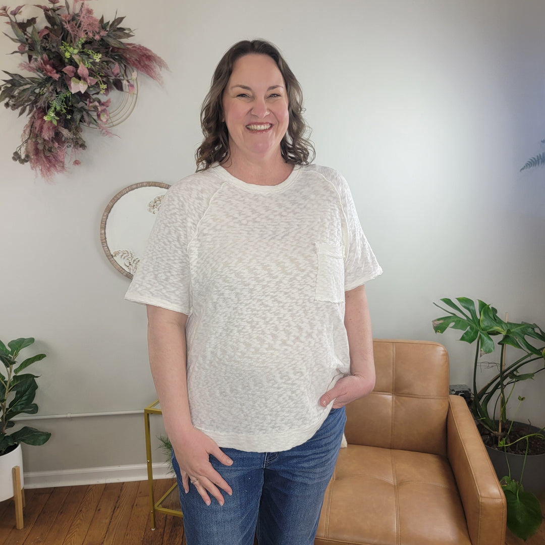 A smiling woman with wavy brown hair stands indoors wearing the Very J Nahla White Slub Tee and blue jeans, next to a tan chair with plants and a floral wall decoration in the background.