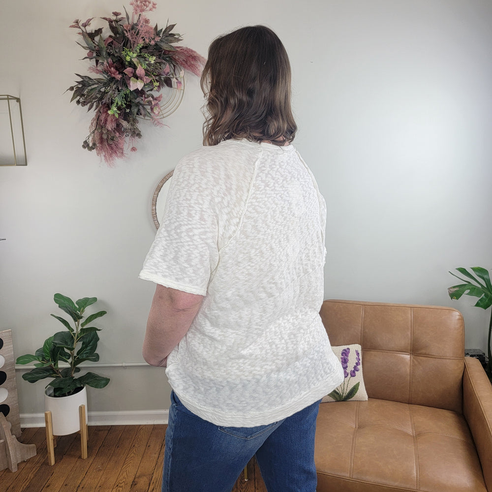 A person with shoulder-length brown hair, wearing the Nahla White Slub Tee by Very J and blue jeans, stands with their back to the camera in a cozy, plant-filled living room with neutral decor.