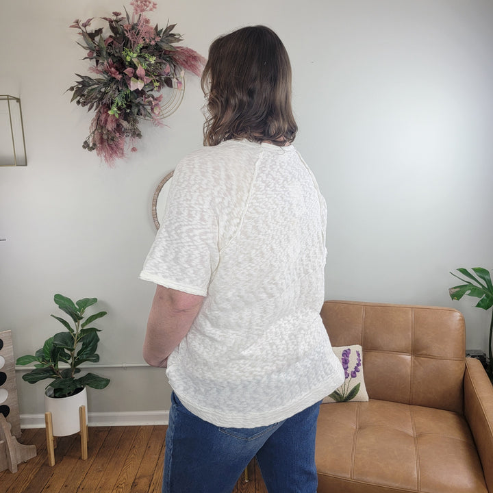 A person with shoulder-length brown hair, wearing the Nahla White Slub Tee by Very J and blue jeans, stands with their back to the camera in a cozy, plant-filled living room with neutral decor.