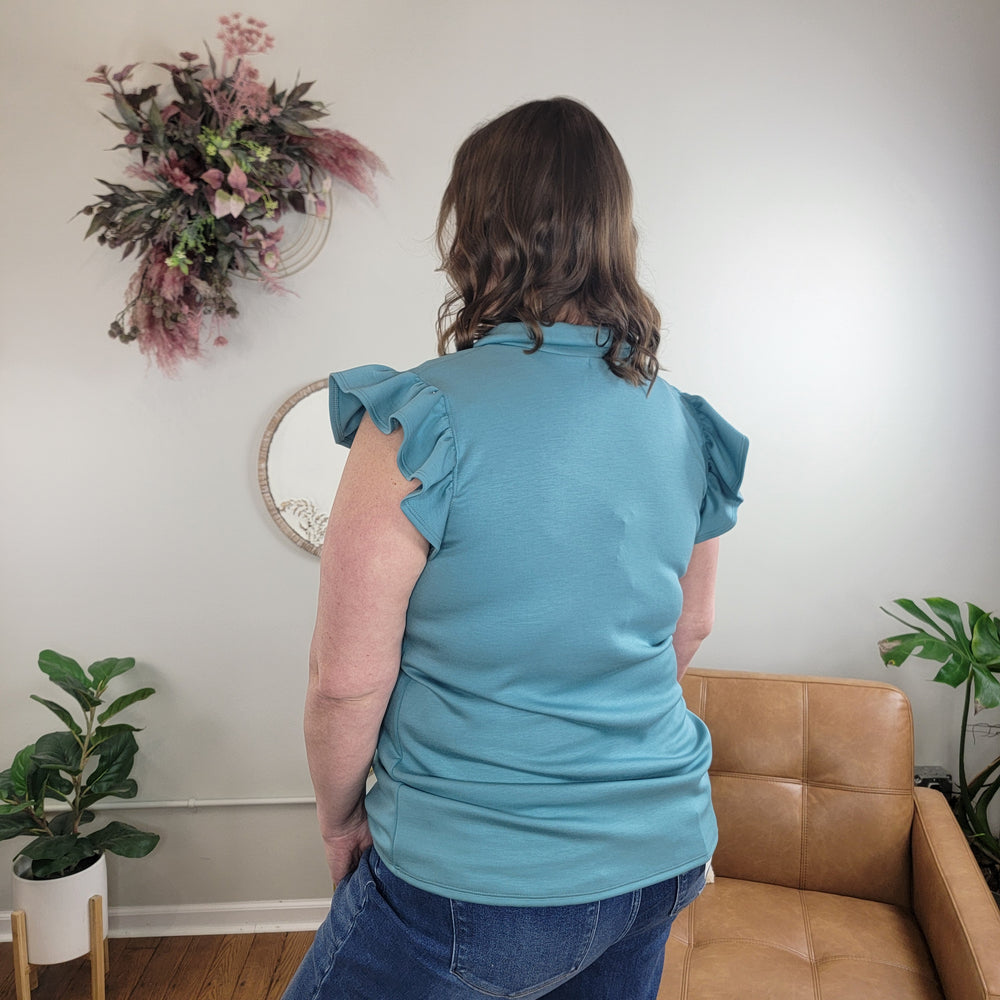 A woman with wavy brown hair, wearing the Heimish Dakotah Ruffle Sleeve Top in teal and jeans, stands with her back to the camera in a cozy room with plants, a tan leather chair, and floral wall decor.
