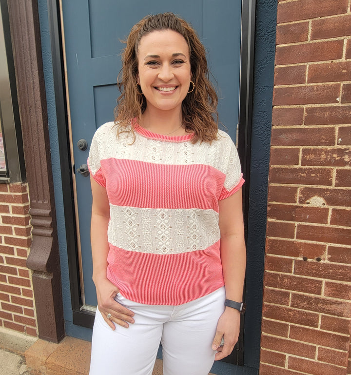 A woman with curly brown hair smiles in front of a blue door and brick wall, wearing the 7th Ray Roxy Lace Detail Top with pink and white stripes and lace, paired with white pants.