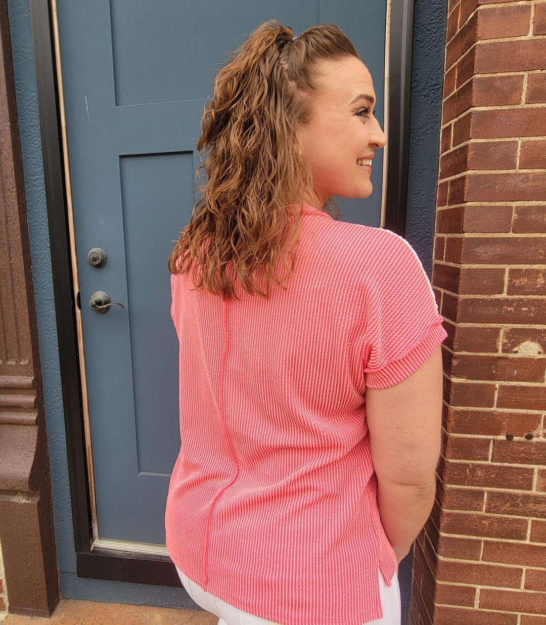 A woman with wavy brown hair tied half-up smiles, facing away by a blue door and brick wall. She wears the 7th Ray Roxy Lace Detail Top in coral and white eyelet lace pants.