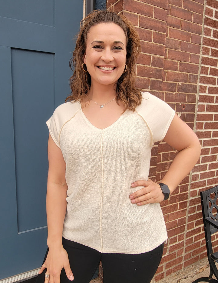 A woman with curly brown hair smiles outside in front of a blue door and red brick wall, wearing the 7th Ray Mia V-Neck Top in cream and black pants, with one hand on her hip.