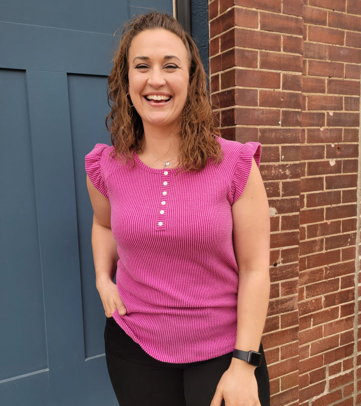 A woman with curly brown hair wears the magenta Mindy Top by 7th Ray and black pants, smiling as she stands in front of a blue door and brick wall.
