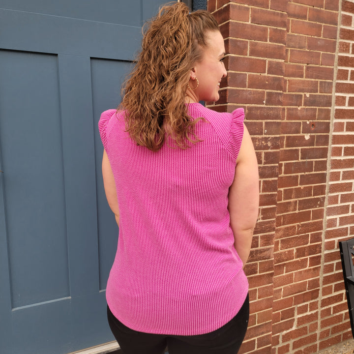 A woman with curly brown hair faces away from the camera, wearing the 7th Ray Mindy Top—a magenta ribbed top with flutter sleeves—and black pants, standing in front of a blue door and brick wall.