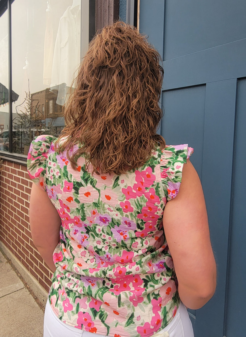 A curly-haired person in front of a blue door wears the Les Amis Marissa Floral Top—pink, red, and green with ruffle sleeves—paired with white pants. City street reflections are visible in a nearby window.