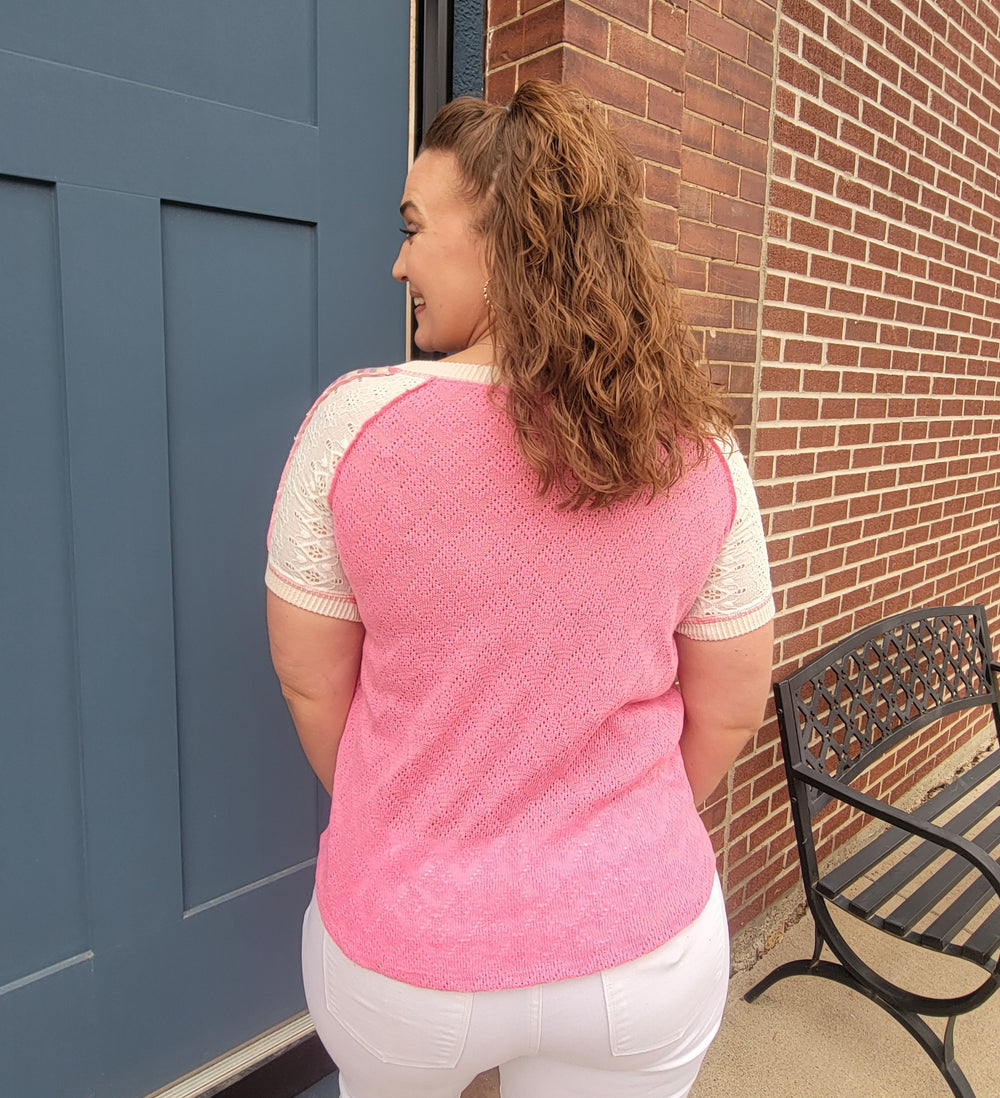 A woman with curly brown hair stands facing away near a blue door and brick wall, wearing the 7th Ray Sloane Patchwork V-Neck Top in pink and white with white pants. A black metal bench is nearby.