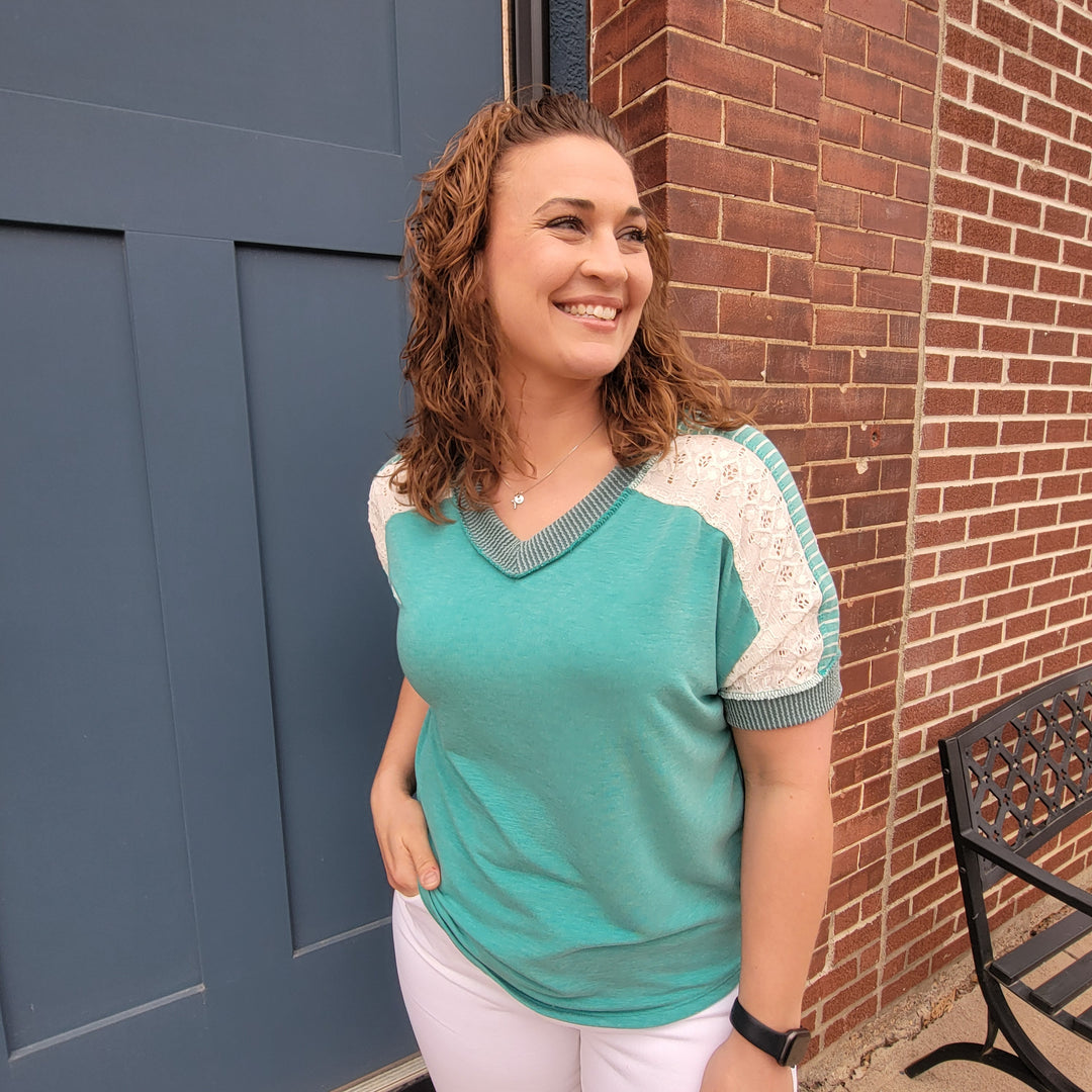 A woman with curly hair stands outside by a blue door and brick wall, smiling in the 7th Ray Lainey Patchwork V-Neck Top with eyelet lace sleeves and white pants, one hand in her pocket as she looks to the side.